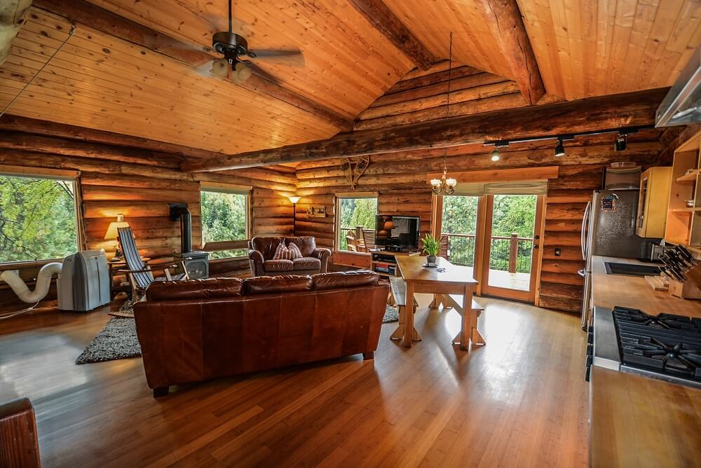 Home remodeling Rustic cabin interior with open-concept living area, natural wood finishes, leather seating, and large windows offering scenic views.