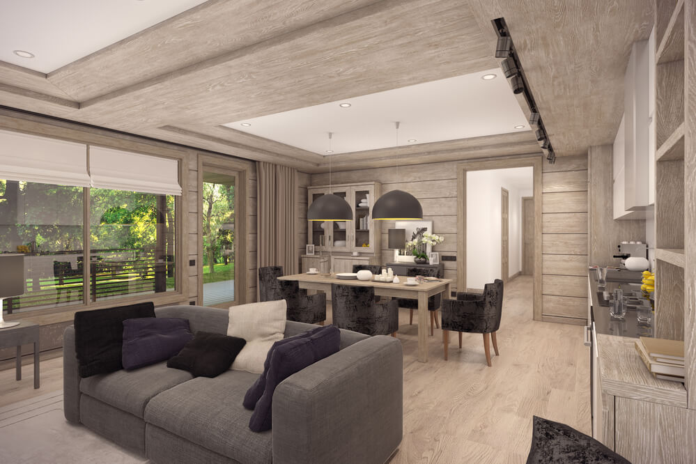 Contemporary home remodeling open-plan living and dining area with a rustic wood finish, featuring a comfortable gray sofa, black accent pillows, a large dining table with upholstered chairs, pendant lighting, and large windows overlooking a green outdoor area.