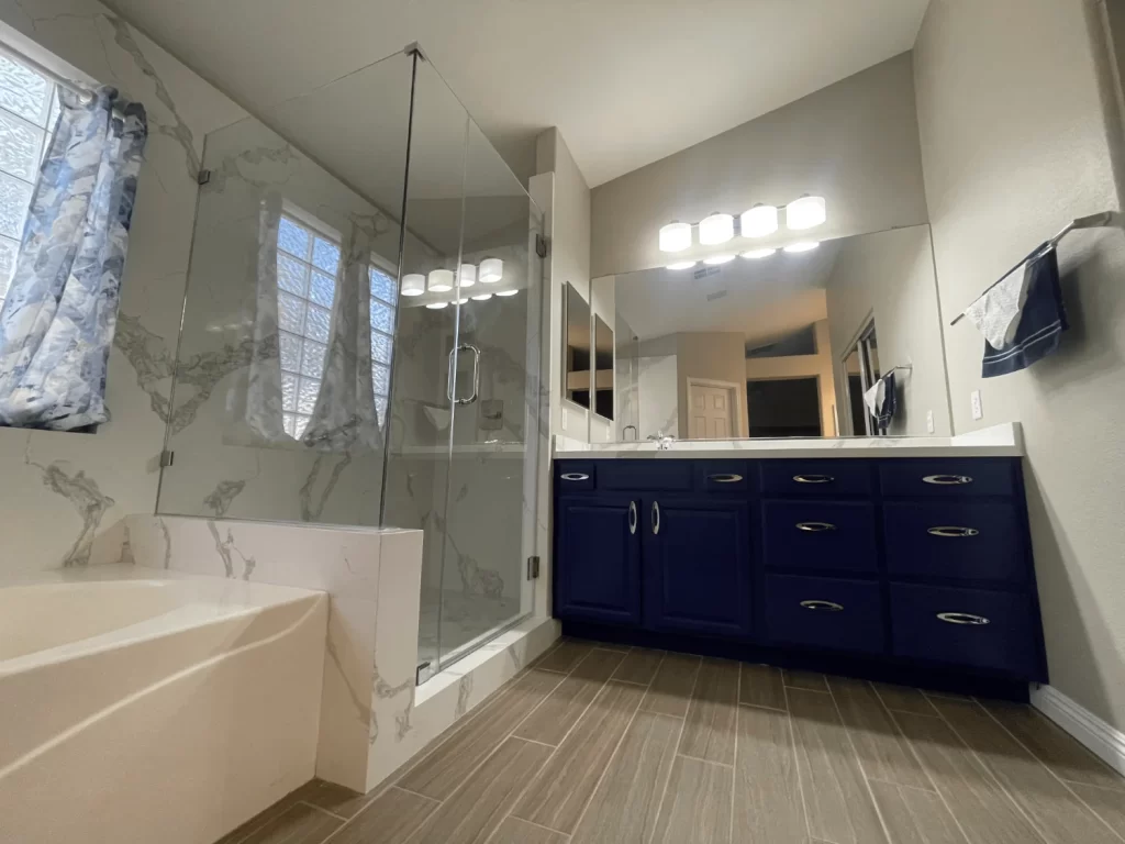 Modern bathroom remodeling design with a navy vanity, glass-enclosed shower, large mirror, and natural tile flooring for a stylish and functional space.