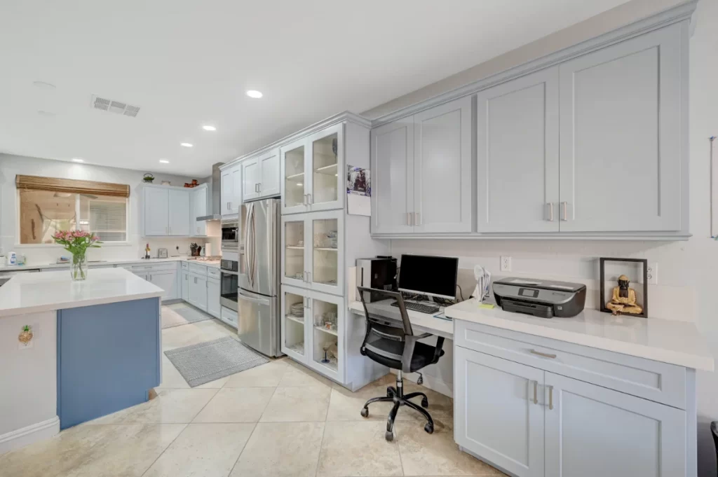 Modern kitchen with light gray cabinets, stainless steel appliances, a built-in workspace with a desk, computer, and printer, and a spacious layout with white countertops and tile flooring.